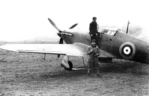 Man in flying suit standing beside single-engined military monoplane; another man stands on the aircraft’s wing, next to the cockpit
