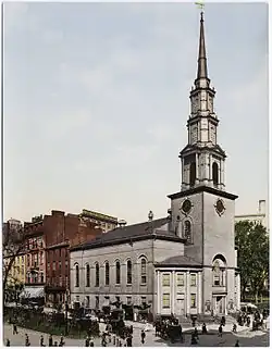 Ground-level view of a brick church with a large, white, tapering spire; a brown skyscraper is visible in the distance, with several shorter high-rises located closer to the church.