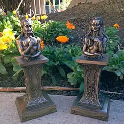 Two brownish metal bust statues on pedestals. On the left, a bearded, aged man with deer horns representing the Horned God. On the right, an aged woman with a waning crescent above her forehead representing the senescent «crone» form of the Triple Goddess.