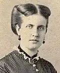 Photographic portrait of a young lady with light-colored hair swept back and wearing a high-necked, Victorian era striped dress, dark earrings and a dark locket suspended around her neck on a ribbon