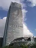 Ground-level view of a tall, mostly glass facade set in front of a blue, cloudy sky; a smaller, circular building is in the foreground